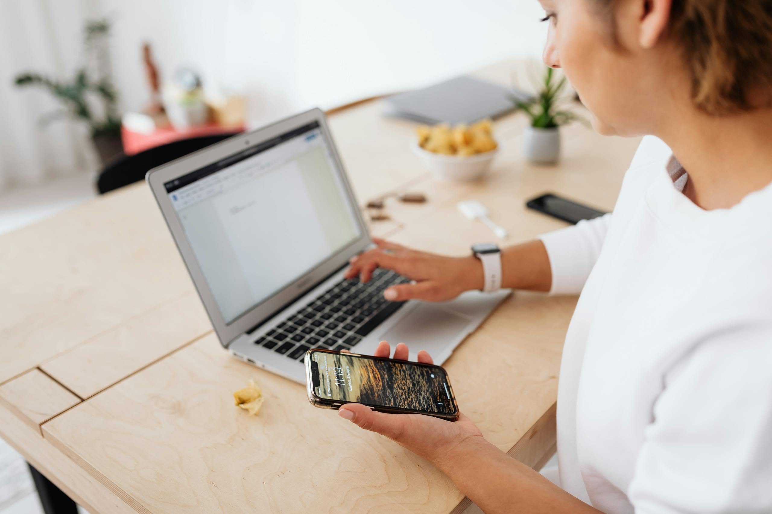 A woman sitting at a wooden table using a laptop and smartphone indoors.