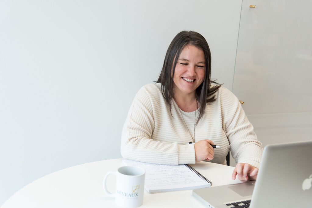 Heather Deveaux sitting at a table against a white background smiling at her laptop