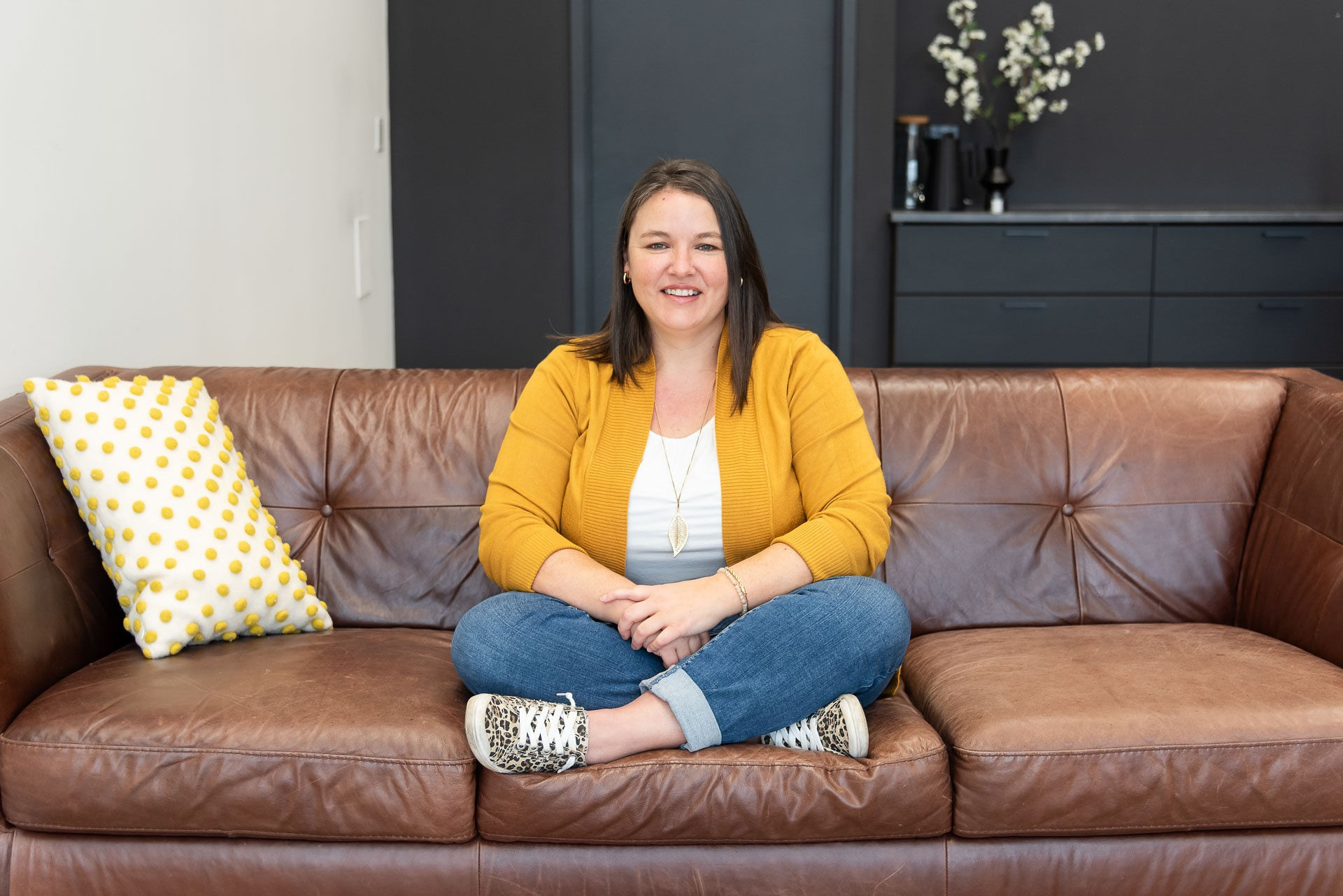 Heather Deveaux sits on a brown leather couch with her legs crossed wearing a yellow cardigan, white shirt and jeans.