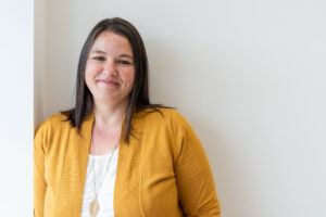 Heather Deveaux is an entrepreneur, author and business coach. She is leaning against a white wall wearing a yellow cardigan