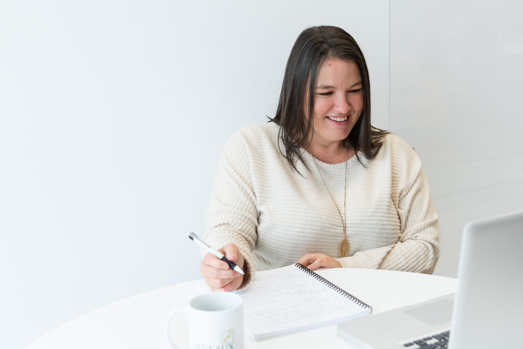 Heather Deveaux sits at a desk smiling at her laptop wearing a white sweater and holding a pen to a notebook.
