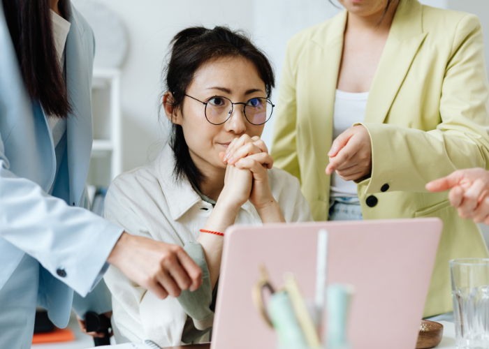 A woman appears tense at her computer while people point out options as she has to make a decision. Heather Deveaux Creative.